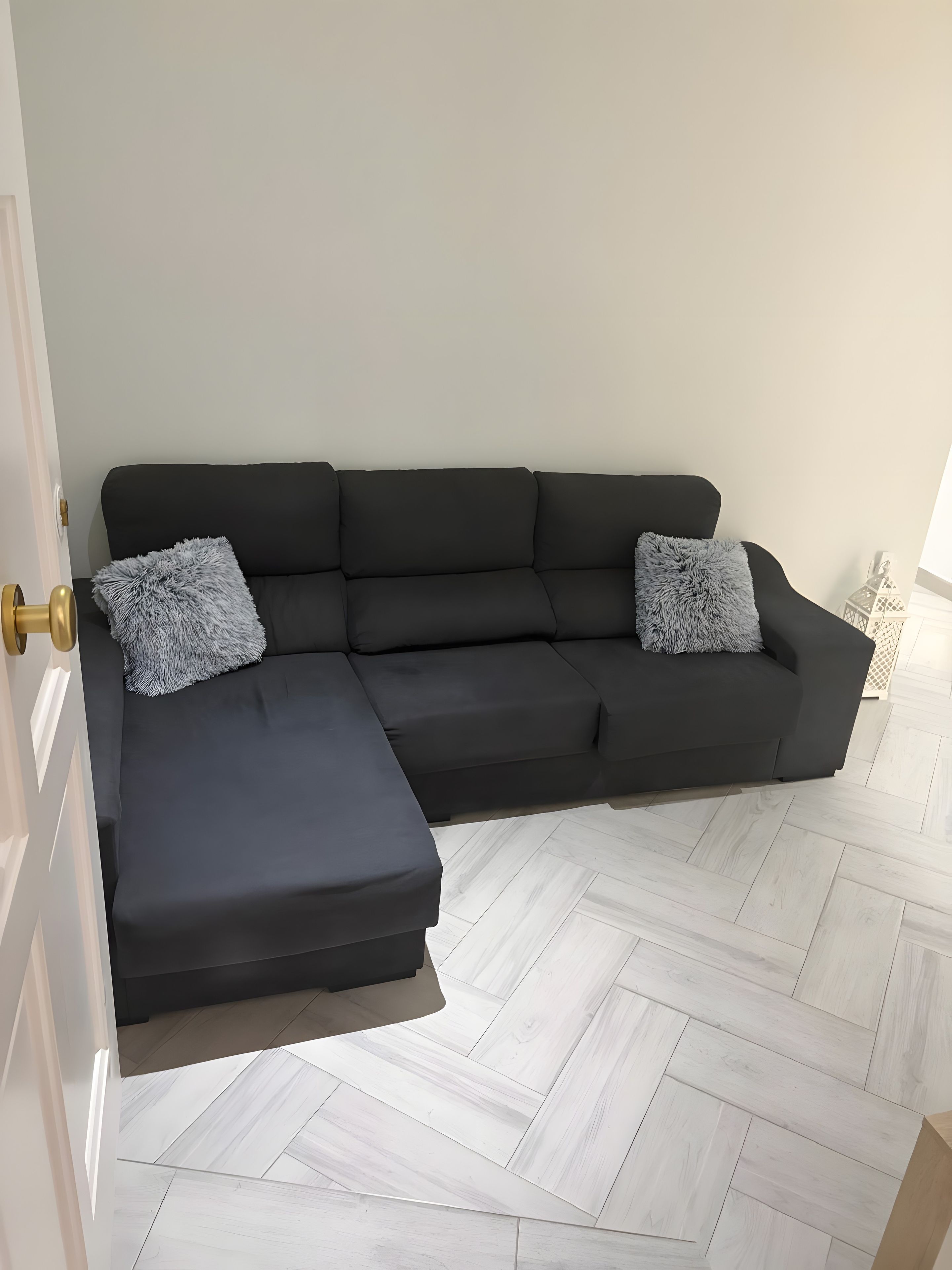 A sectional sofa with two grey pillows, a door with a brass handle, and a herringbone-patterned floor.