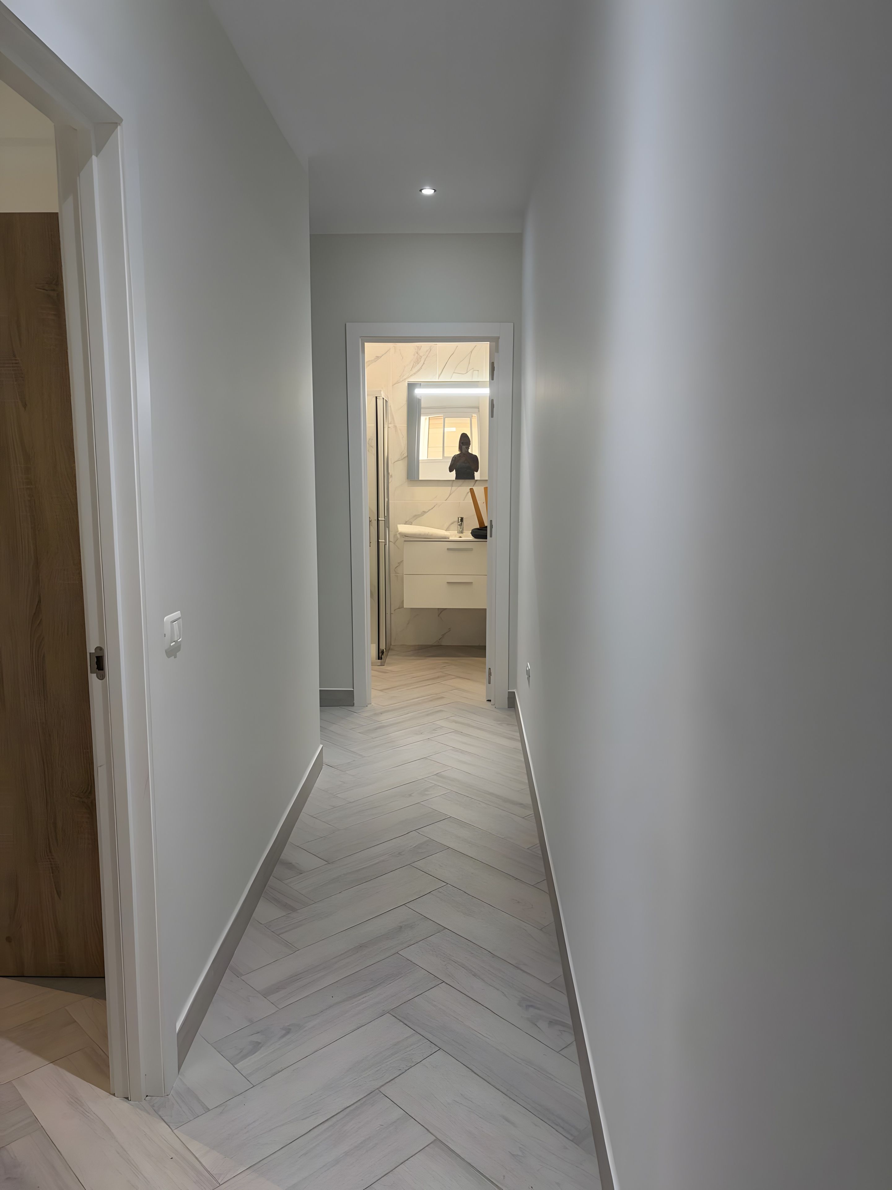 A hallway with a herringbone floor pattern leading to a bathroom with a mirror, sink, and toilet.
