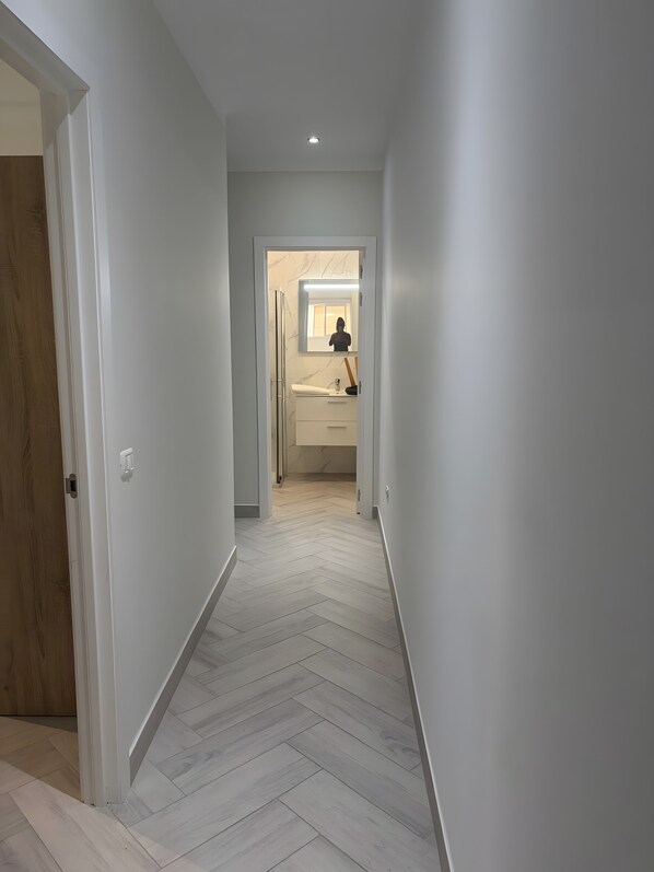 Un couloir dont le sol est recouvert d’un parquet à motifs en chevrons, menant à une salle de bain équipée d’un miroir, d’un lavabo et de toilettes.