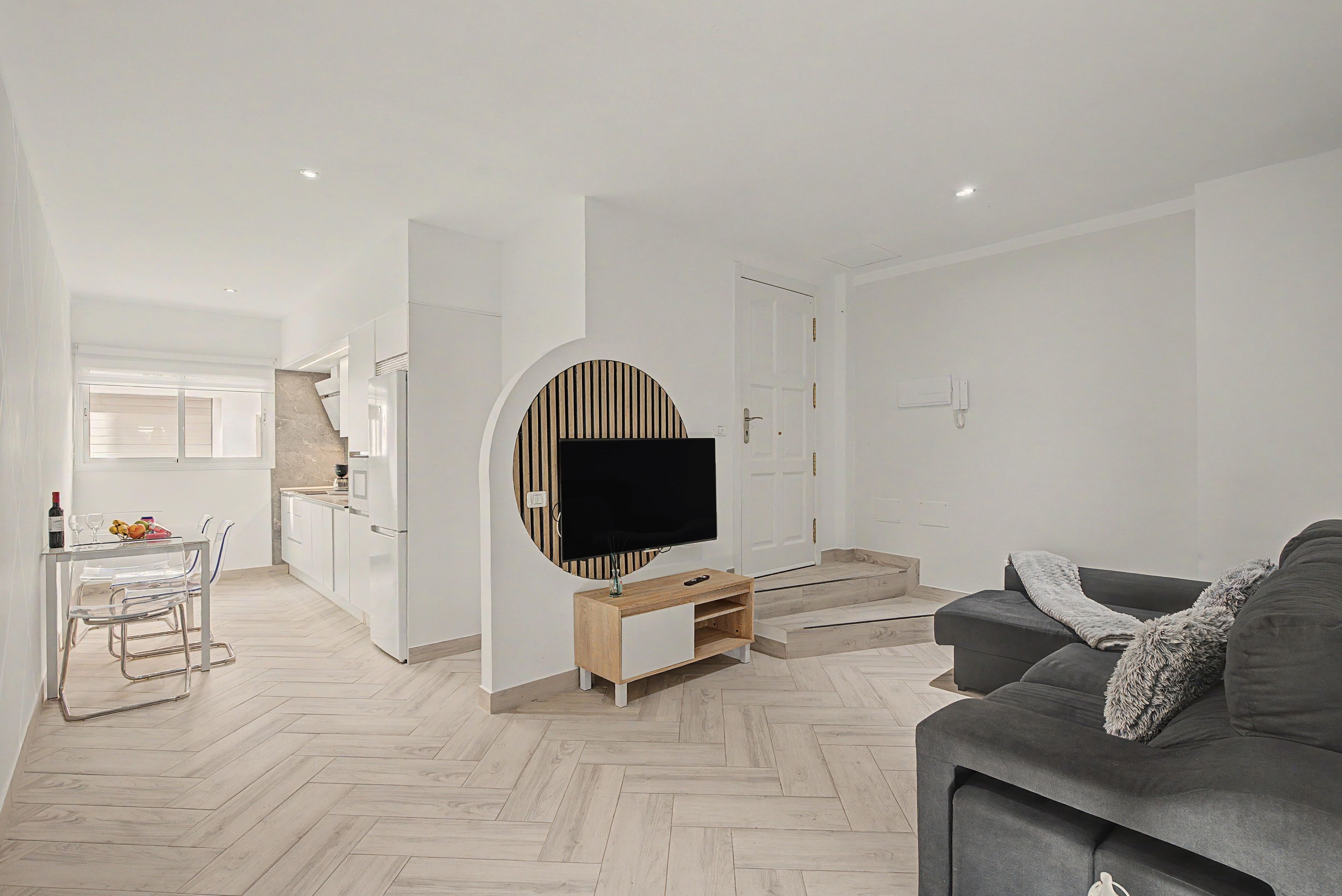 A modern living room with a grey sofa, a flat-screen TV mounted on a wall, and a herringbone-patterned wooden floor.