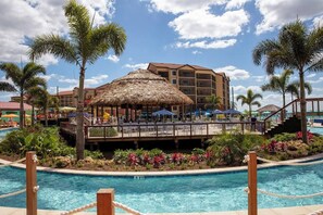 A resort pool area with a thatched-roof structure, palm trees, and a multi-story building in the background. - Studio Villa Westgate Near Disney and Universal (Orlando)