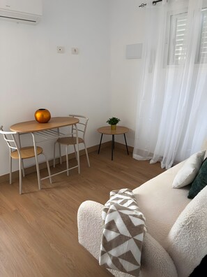 Un espacio habitable moderno con sofá, mesa de comedor con sillas, una mesita auxiliar con una planta y una ventana con cortinas translúcidas.