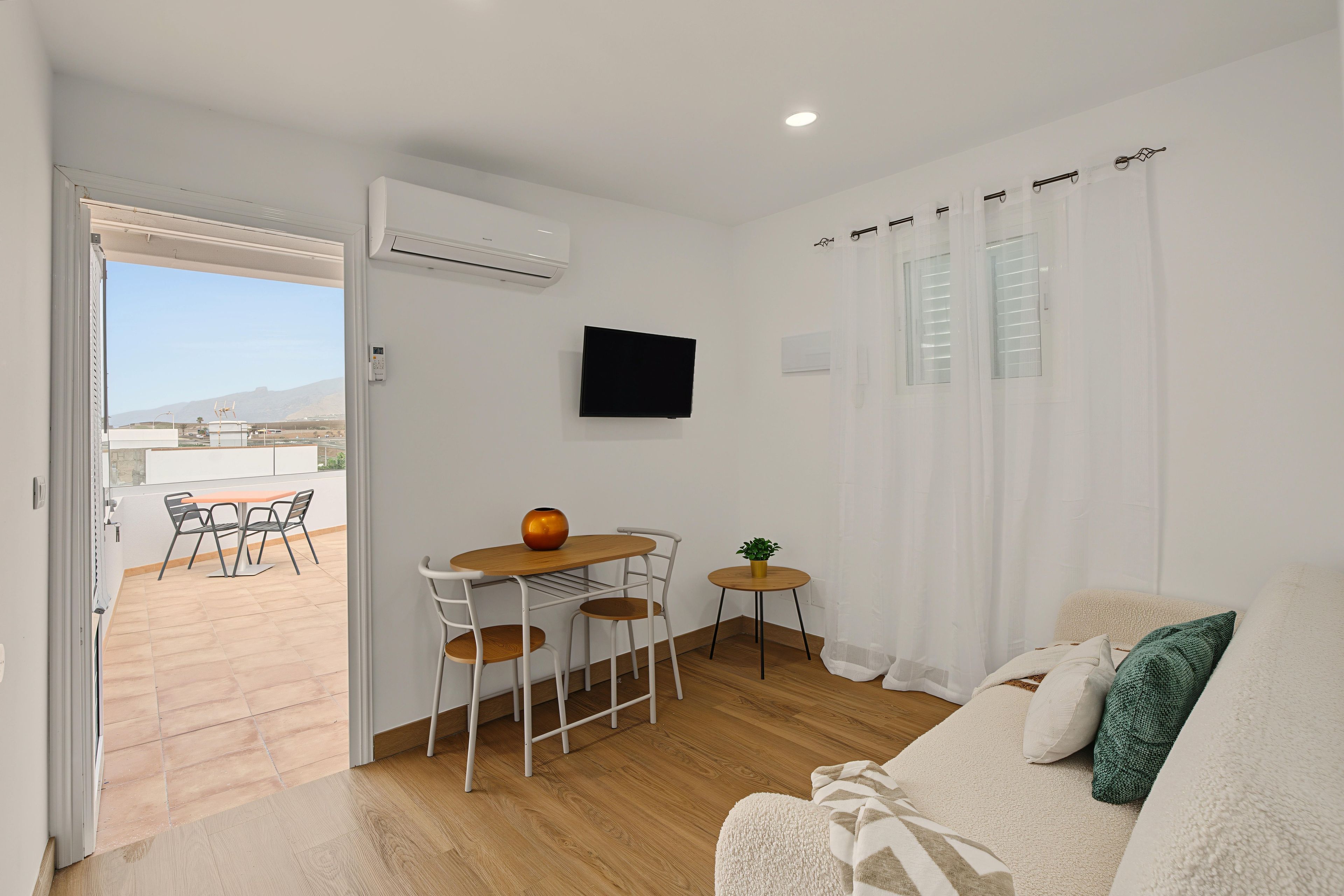 A living room with a sofa, a TV mounted on the wall, a dining area with a table and chairs, and an air conditioning unit.