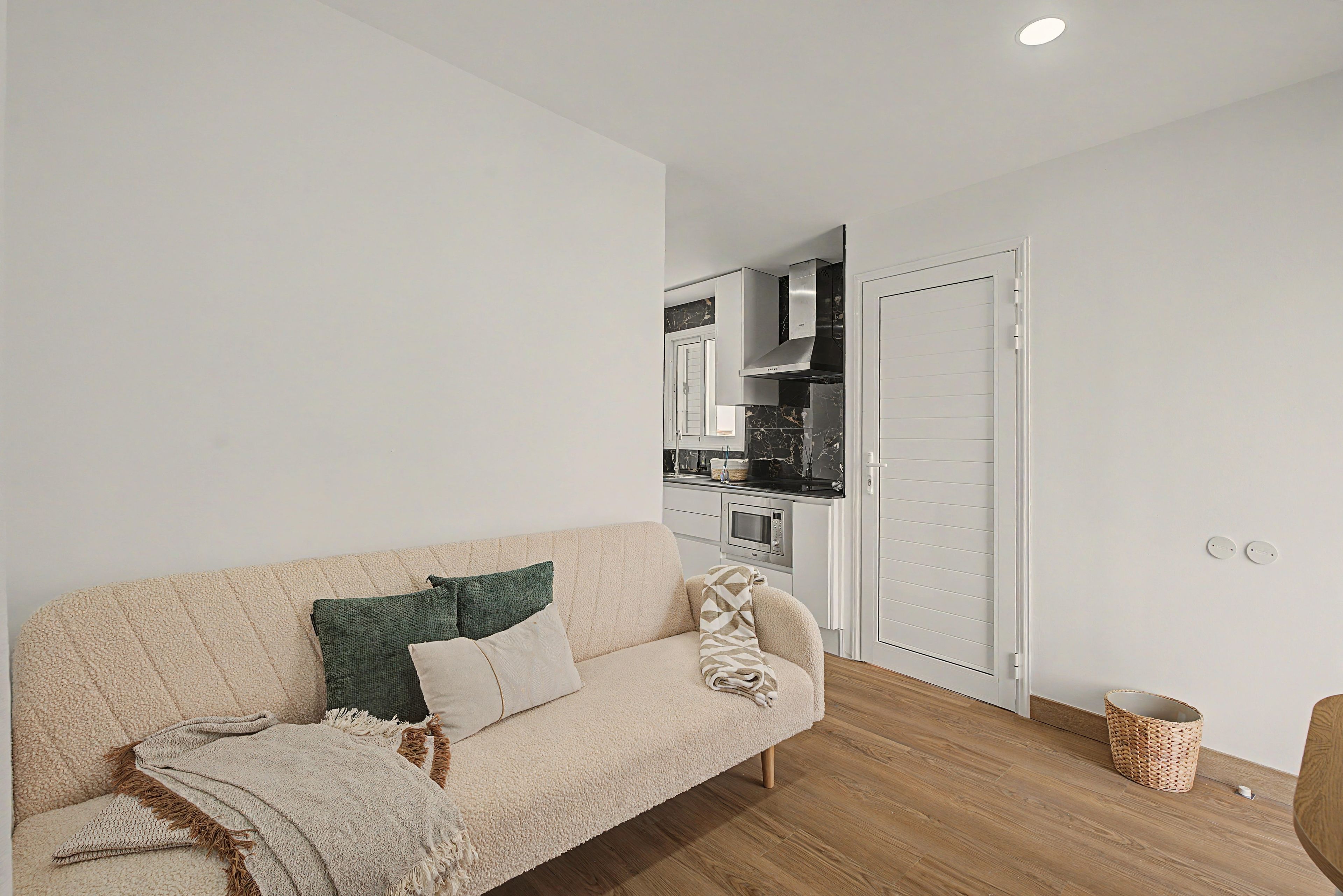A living room with a sofa, a kitchen in the background, and a wooden floor.