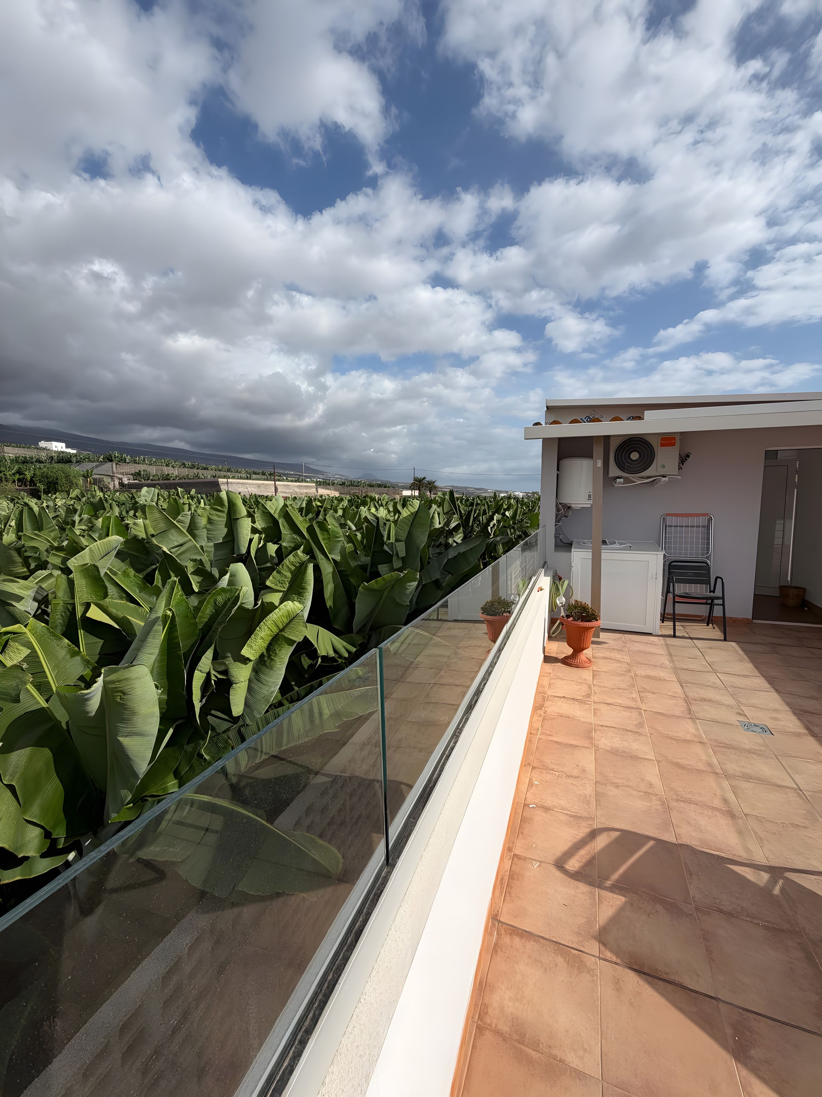 Un balcón con barandilla de vidrio que da a un jardín, un pequeño espacio de estar con una silla y una mesa, y vista al cielo nublado.