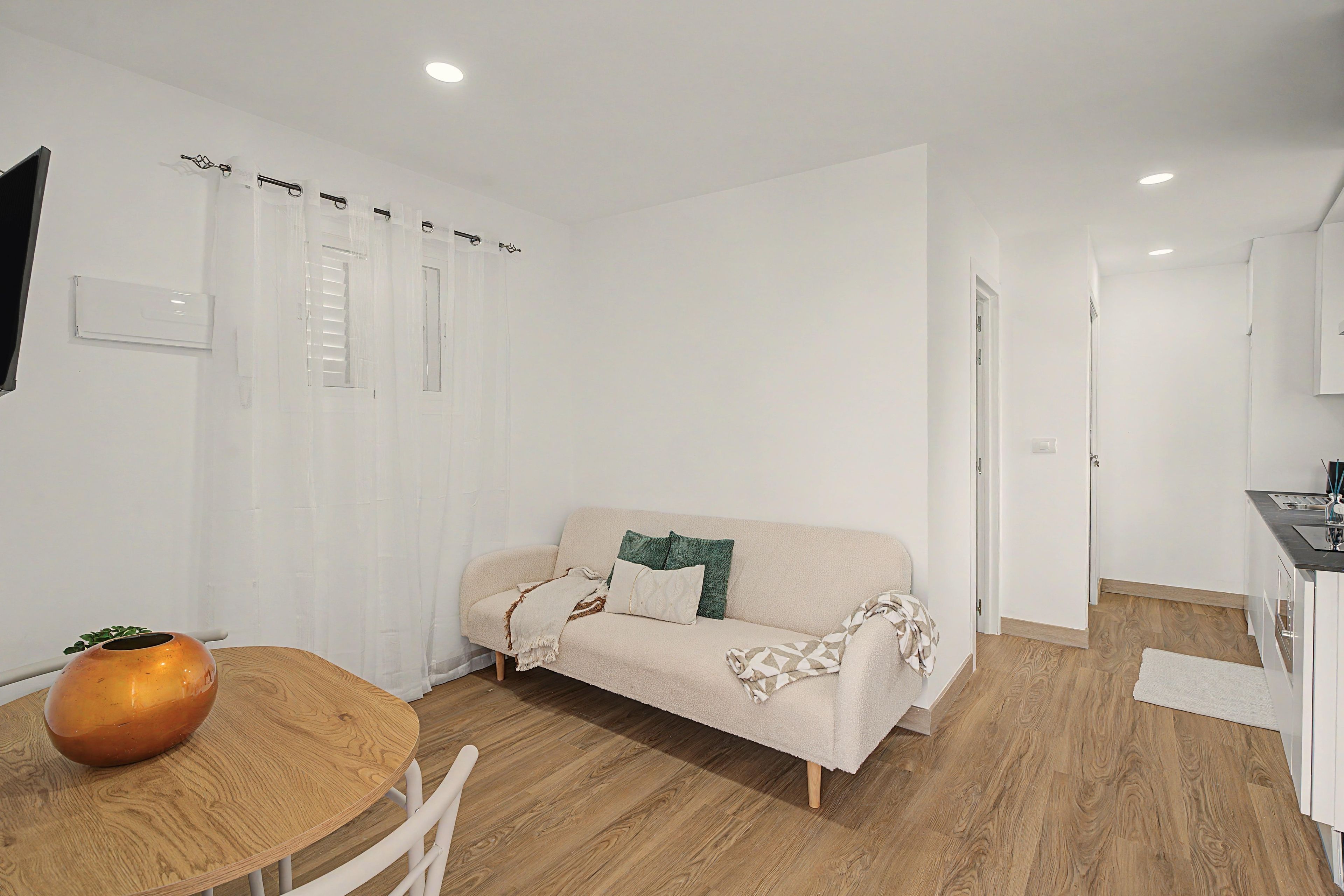 A modern, minimalistic living space with a sofa, a round wooden table, and a kitchen area.