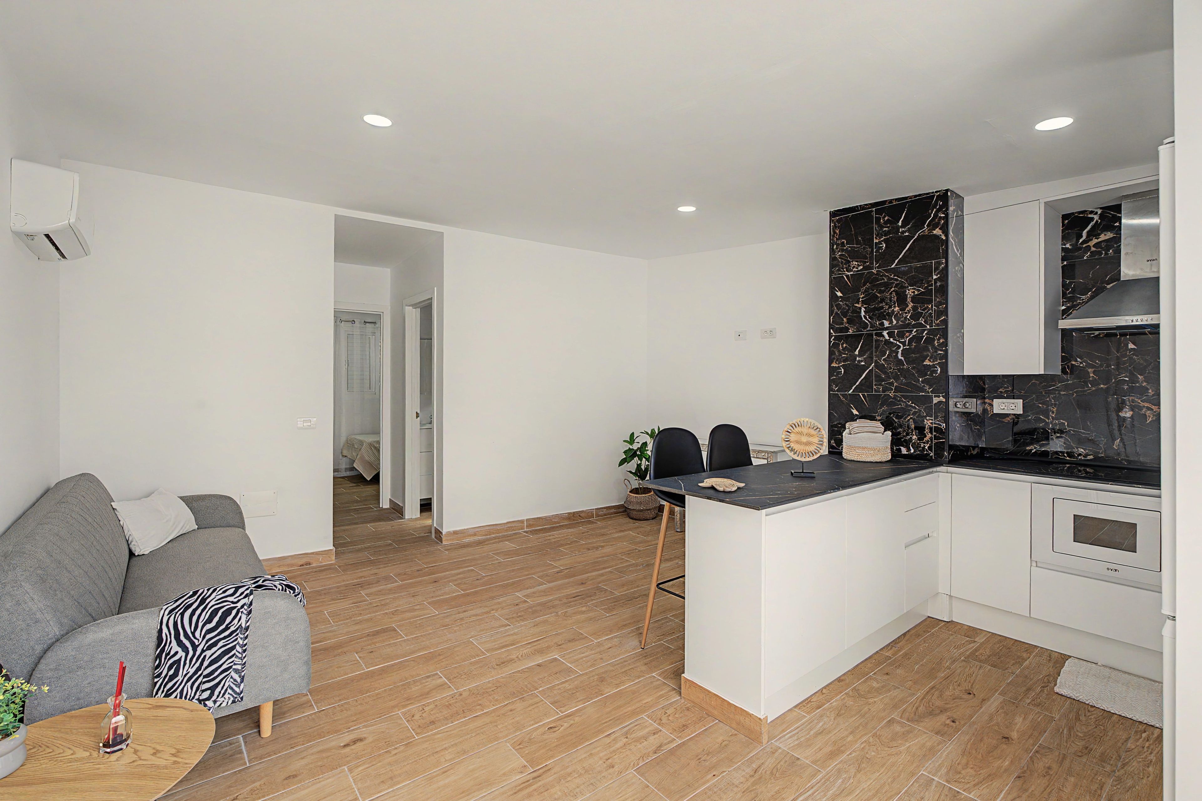 Un apartamento moderno con sofá gris, una pequeña mesa de comedor, una cocina con salpicadero de mármol negro y suelo de madera.
