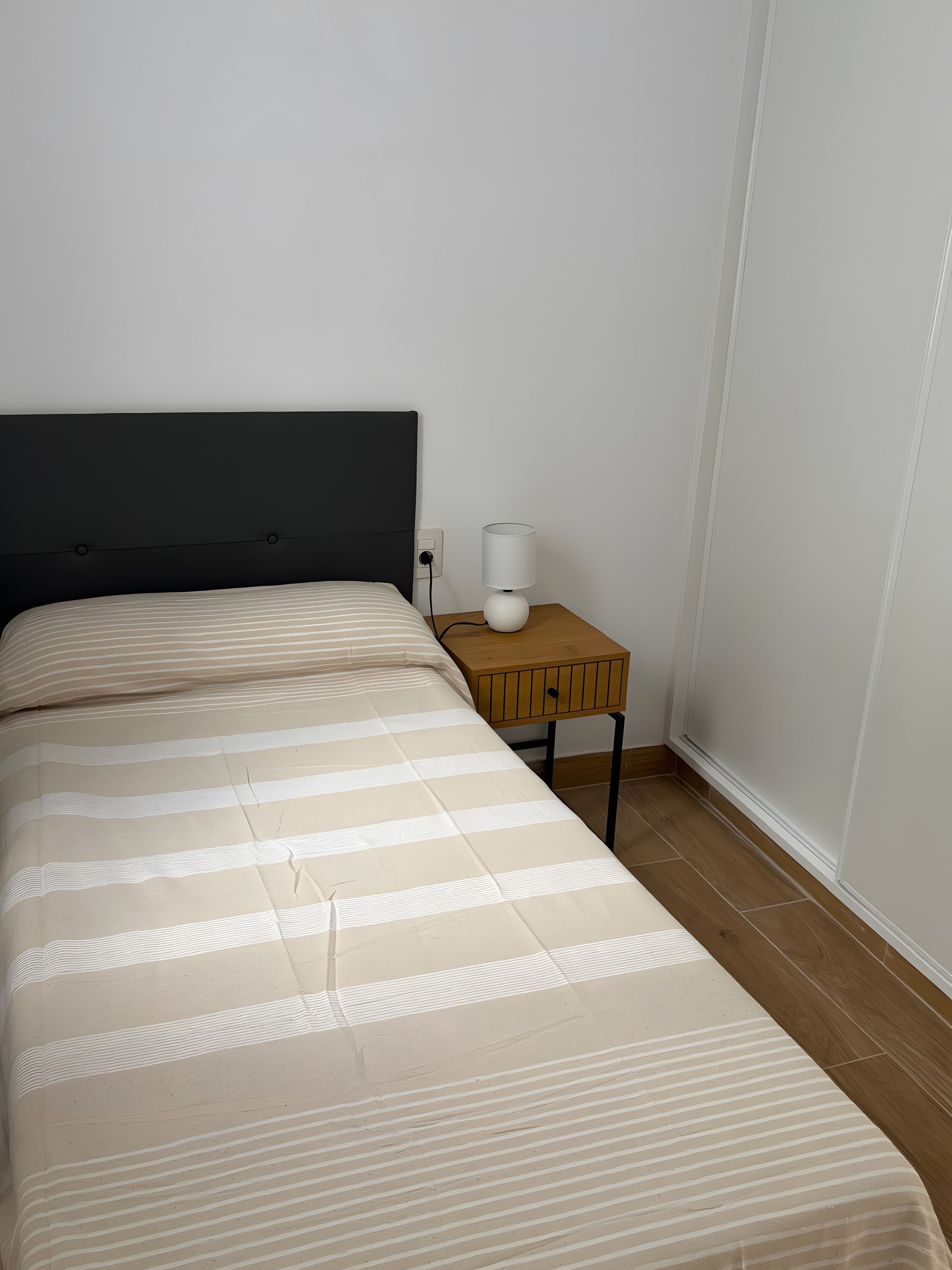 Una cama con colcha a rayas, una mesita de noche con lámpara y un enchufe de pared.