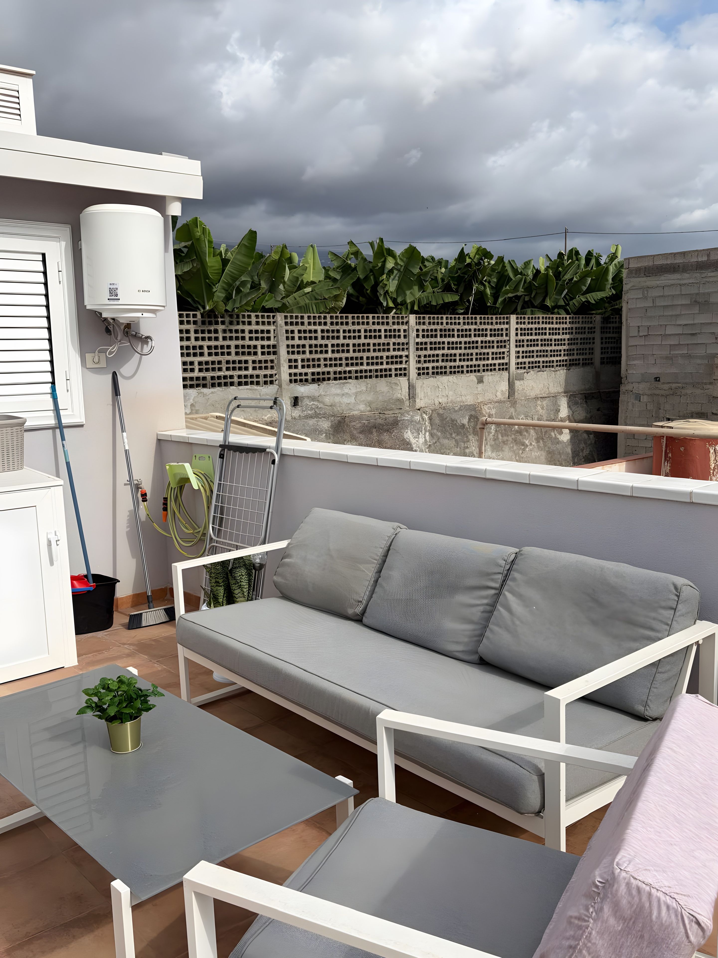 Ein Balkon mit einer grauen Couch, einem kleinen Tisch mit einer Pflanze, einem Besen und einem Warmwasserbereiter.