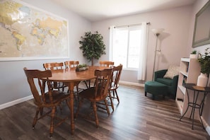 A dining room with a wooden table and chairs, a world map on the wall, a potted plant, a green armchair, and a floor lamp.