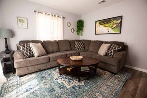 A living room with a sectional sofa, a round coffee table, a patterned rug, a lamp, and wall art.