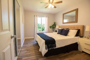 A bedroom with a bed, bedside tables, a ceiling fan, a window with curtains, and a plant.
