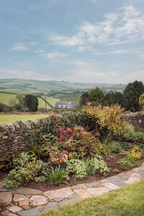 A stone wall with a garden in the foreground, a stone building in the middle ground, and rolling hills in the background. - Penlows (Blackawton)