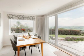 A dining room with a wooden table set for a meal, large windows offering a view of the countryside, and a view of the sky. - Penlows (Blackawton)