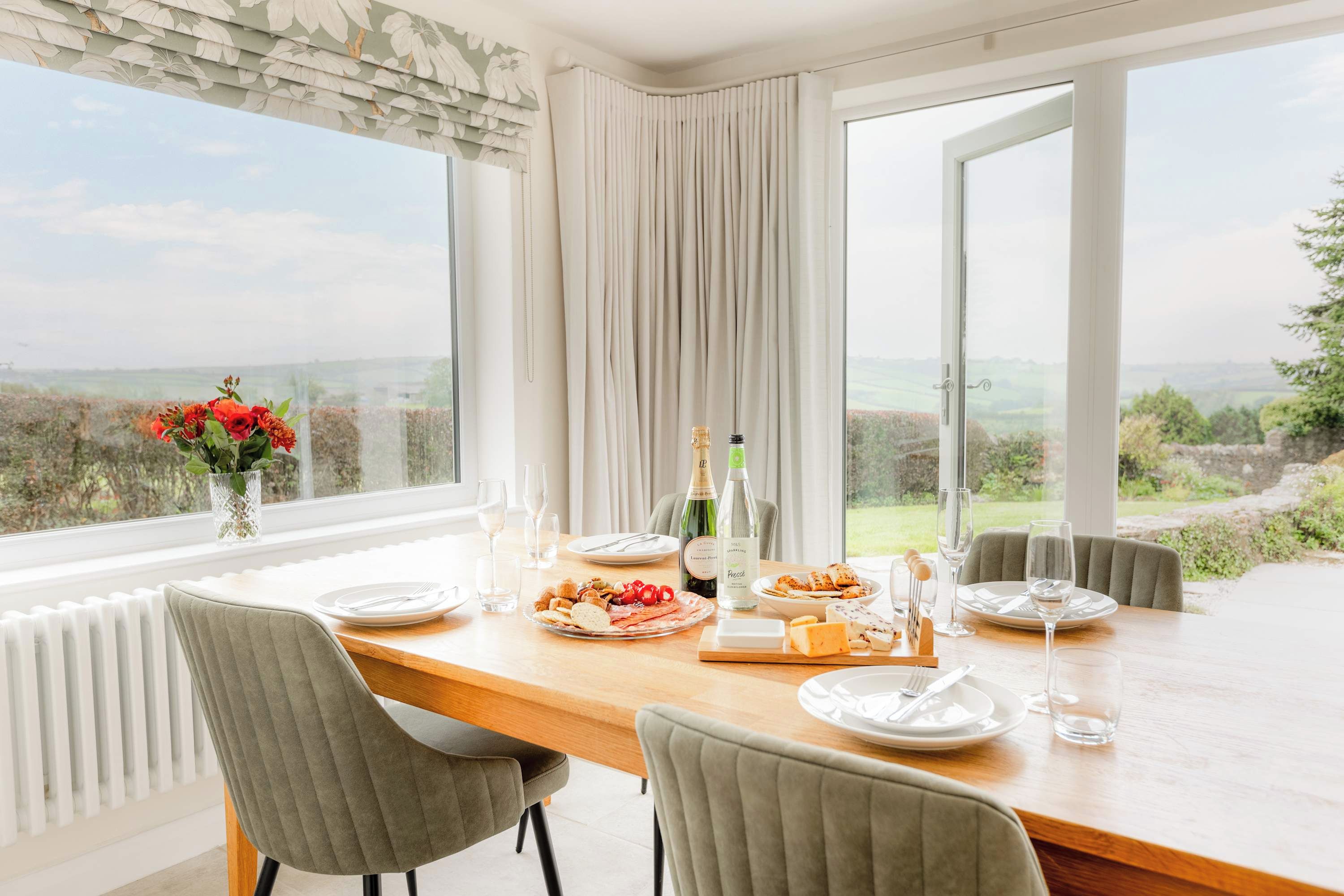 A dining table with a view of the countryside, set with plates, glasses, and a bottle of champagne.
