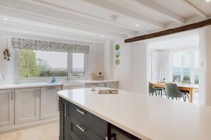 A modern kitchen with a large island, a dining area with a table and chairs, and a view of the outdoors through large windows. - Penlows (Blackawton)