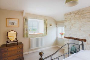 A bedroom with a stone wall, a dresser, a radiator, a window with curtains, and a bed with a metal headboard. - Penlows (Blackawton)