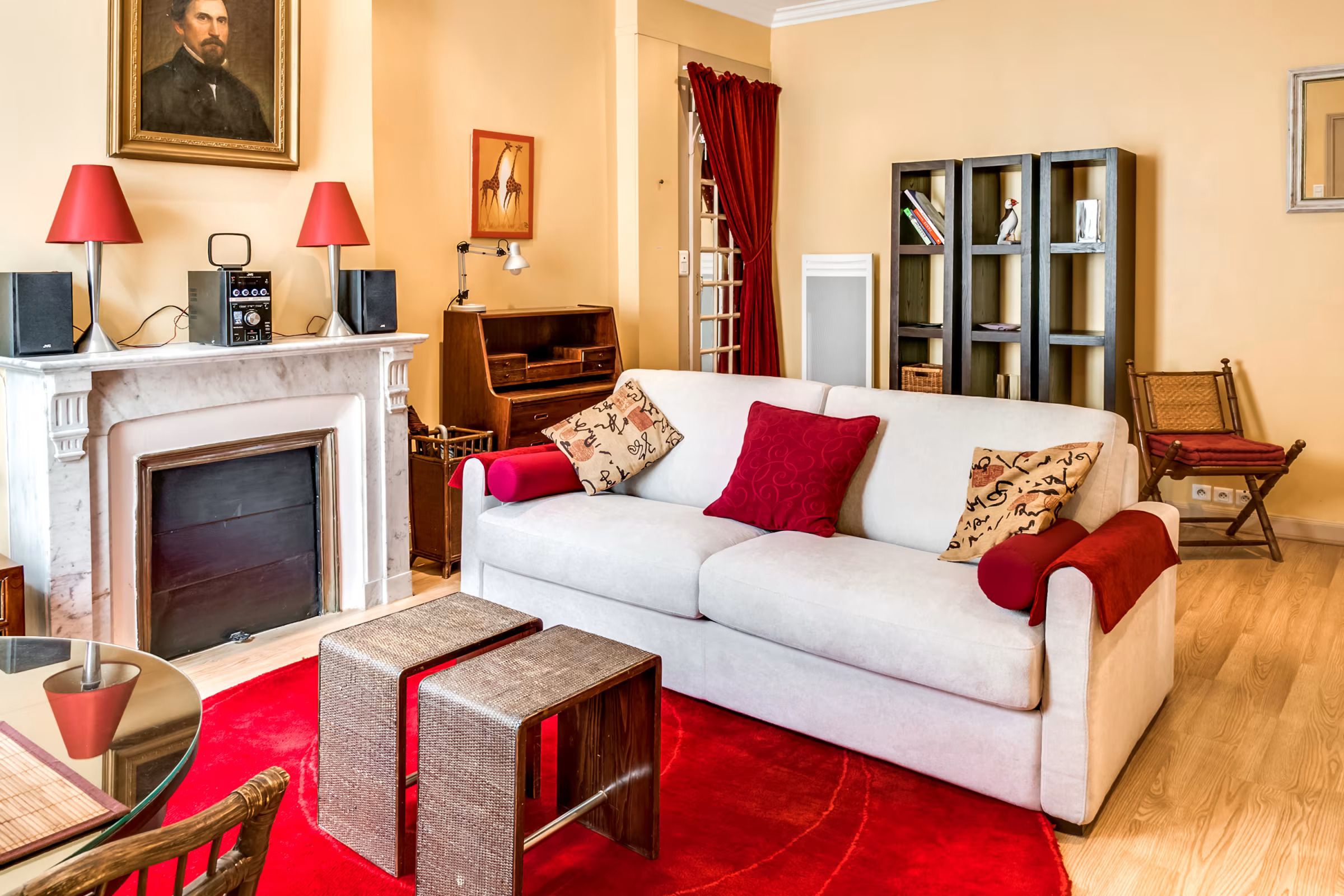 Een woonkamer met een witte bank, rode kussens, een open haard, een boekenkast en een rood vloerkleed.