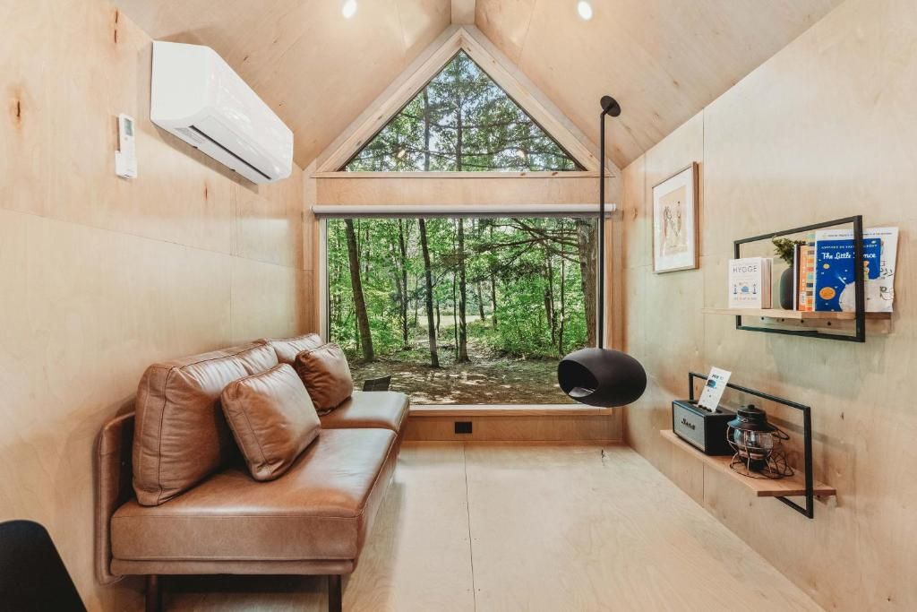 A cozy cabin interior with a wooden ceiling, a leather sofa, a large window with a view of a forest, and a wall-mounted shelf with books and a lantern.