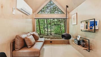 A cozy cabin interior with a wooden ceiling, a leather sofa, a large window with a view of a forest, and a wall-mounted shelf with books and a lantern.