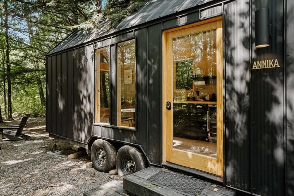 A small, black metal building with a wooden door and windows, labeled 'ANNIKA,' in a wooded area with a picnic table nearby. - Lumen Nature Retreat (Woodstock)