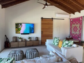 Ein Wohnzimmer mit einem weißen Sofa, einem an der Wand montierten Fernseher, einem Deckenventilator und einer Holztür.