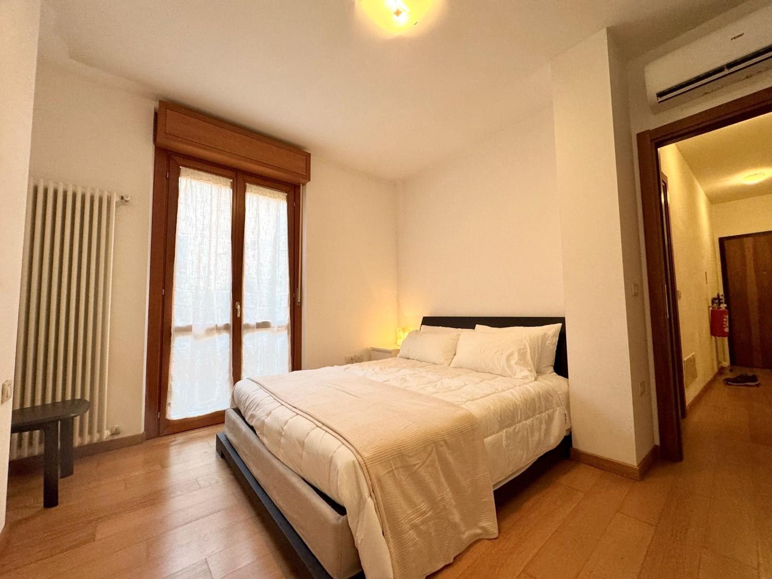 A bedroom with a bed, a radiator, a bench, and a door leading to another room.