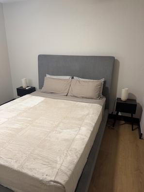 Un lit bien fait, avec une tête de lit grise, deux tables de chevet avec des lampes et un plancher en bois.