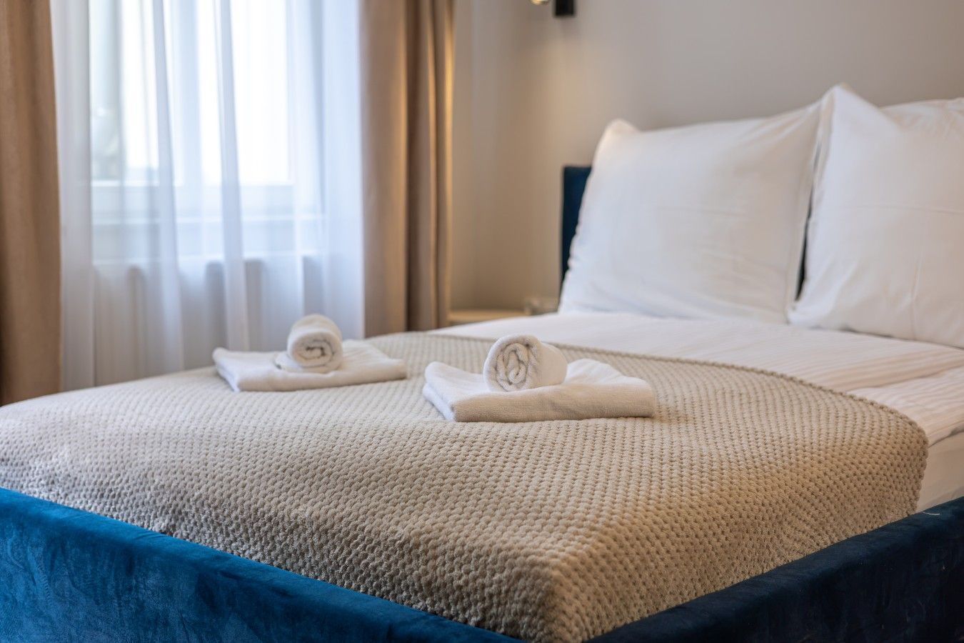 Una cama bien hecha con sábanas blancas y una colcha azul, una toalla enrollada y un almohadón sobre la cama.