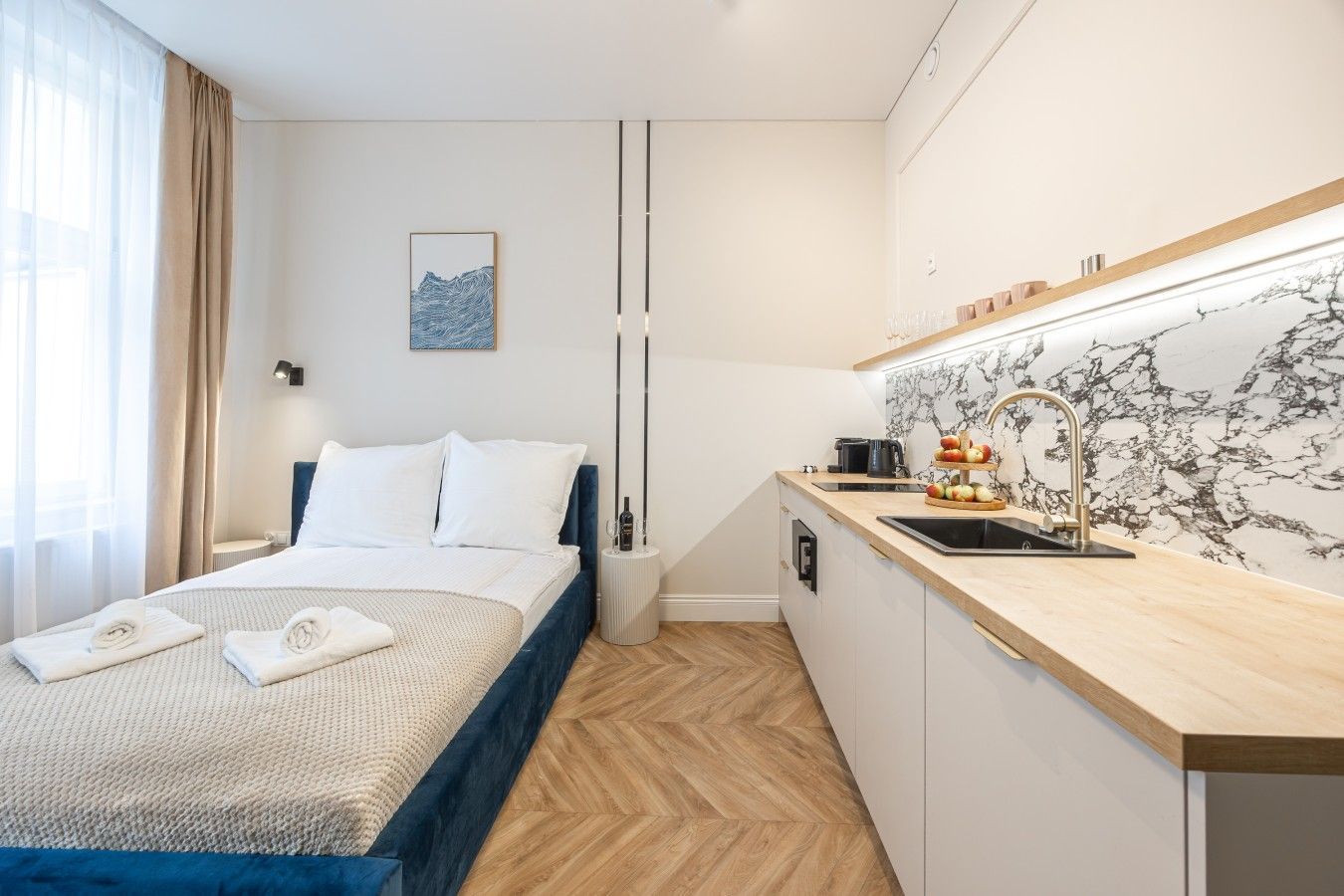 A hotel room with a bed, a bedside table, a small kitchen area with a sink, and a marble backsplash.