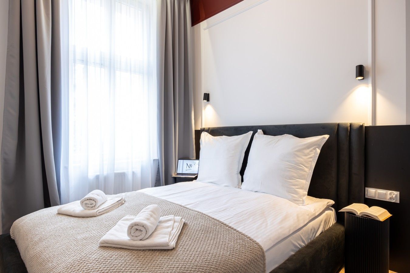 A hotel room with a bed, a bedside table with a lamp, a nightstand with a book, and a window with curtains.