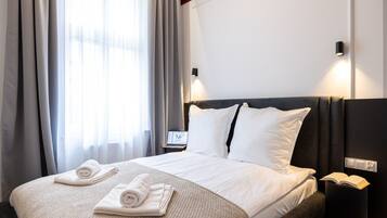 A hotel room with a bed, a bedside table with a lamp, a nightstand with a book, and a window with curtains.
