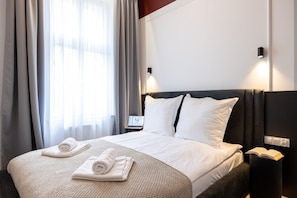 Quarto de hotel com uma cama, mesa de cabeceira com abajur, criado-mudo com um livro e janela com cortinas.