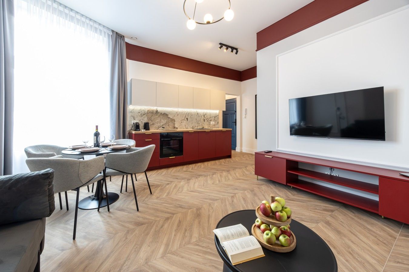A modern living room with a flat-screen TV, a dining area with a table and chairs, and a kitchen with red cabinetry.