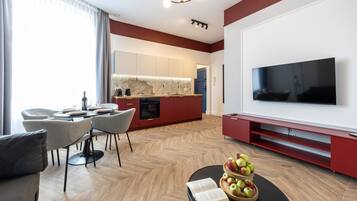 A modern living room with a flat-screen TV, a dining area with a table and chairs, and a kitchen with red cabinetry.