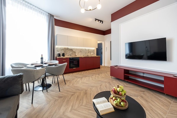 Uma sala de estar moderna com televisão de tela plana, uma área de jantar com mesa e cadeiras, e uma cozinha com armários vermelhos.