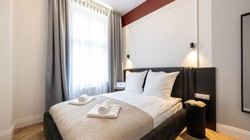 A hotel room with a large bed, a bedside table, a nightstand, a lamp, and a window with curtains.