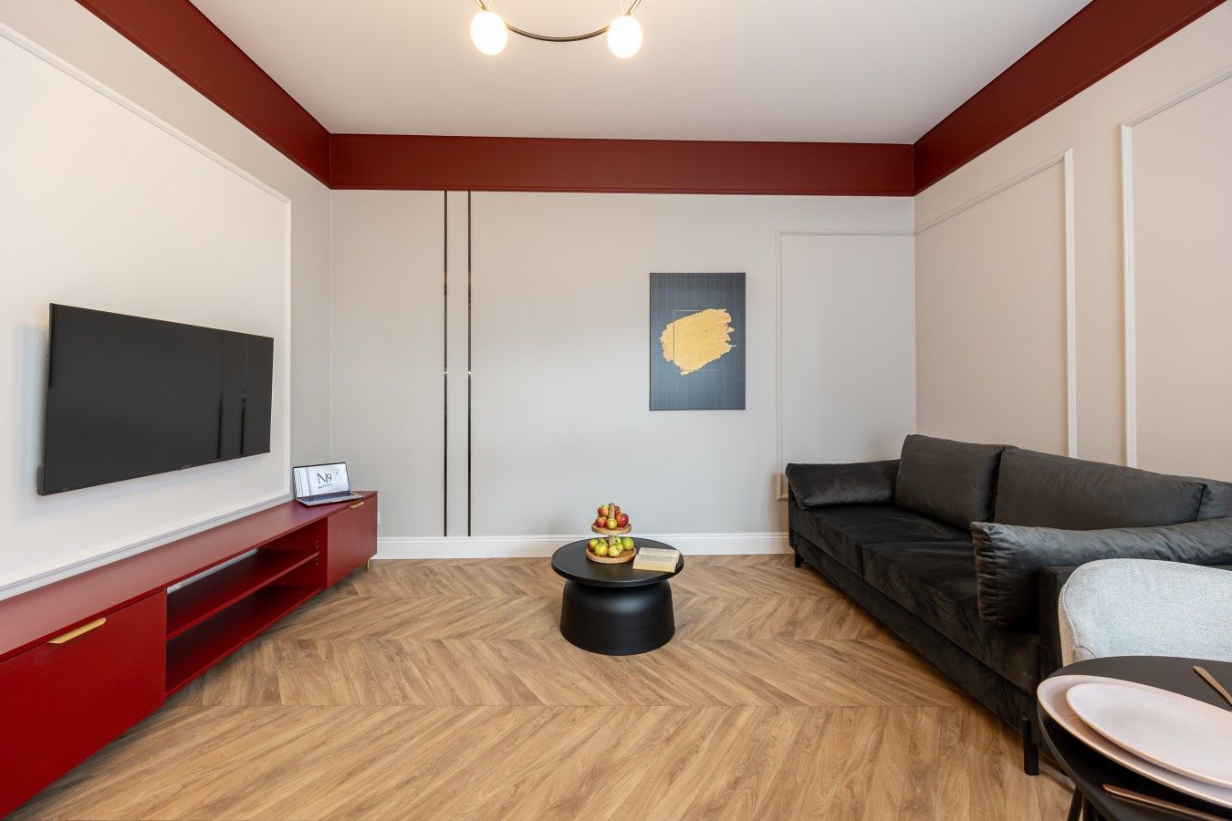 A modern living room with a flat-screen TV, a red entertainment unit, a black sofa, and a round coffee table with fruit.