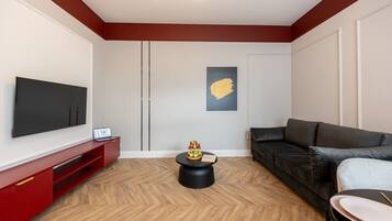 A modern living room with a flat-screen TV, a red entertainment unit, a black sofa, and a round coffee table with fruit.