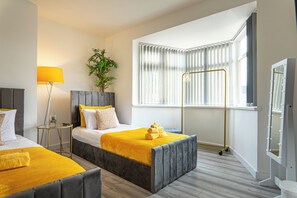 Quarto de hotel com duas camas, uma luminária de chão, um vaso de plantas, uma janela com persianas e uma mesa de toucador com iluminação.