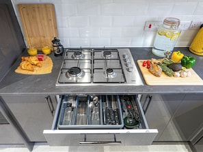 Una cocina con estufa a gas, tabla de cortar con alimentos y cajón con utensilios.