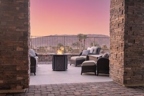 An outdoor seating area with wicker furniture and a central fire feature, surrounded by stone walls and a view of distant mountains. - Lake Las Vegas Home (Henderson)