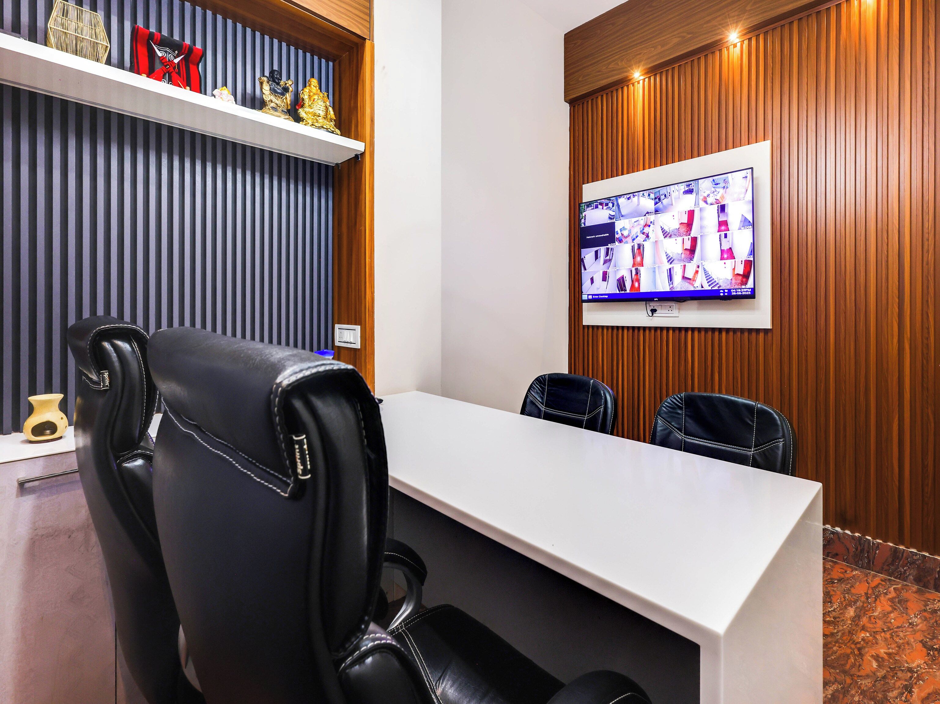 Un salón de conferencias con una mesa blanca, sillas de cuero negro, un televisor montado que muestra imágenes de seguridad y objetos decorativos en estanterías.