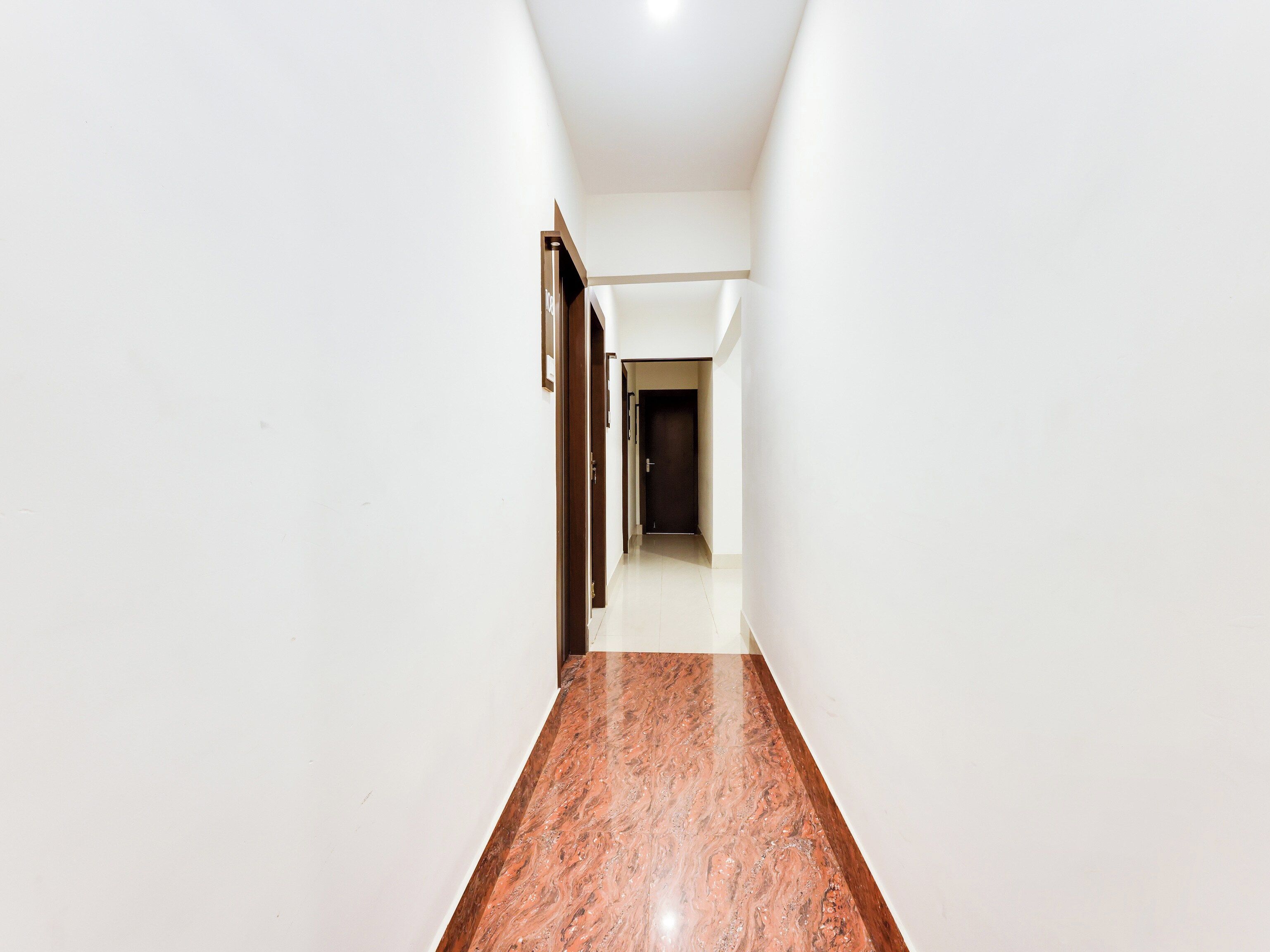 Un pasillo con paredes blancas y un piso de mármol pulido que conduce a una puerta oscura al final.