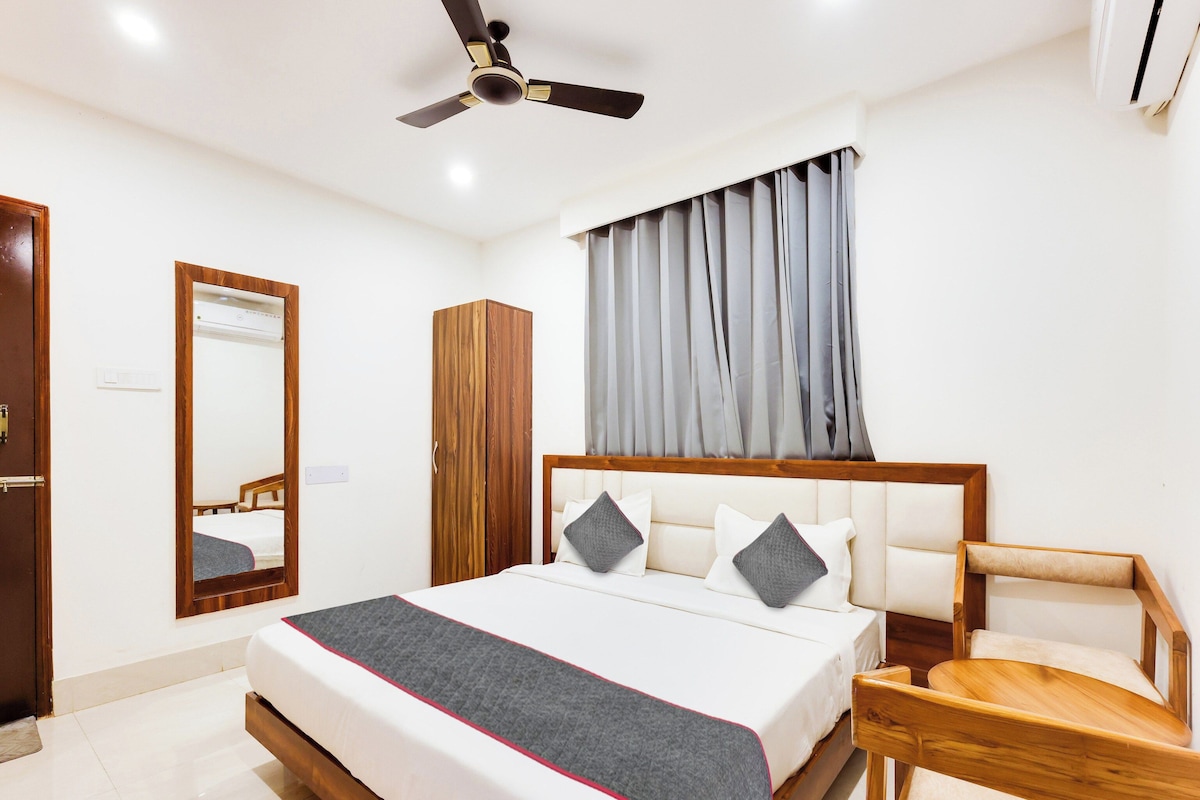 A hotel room with a bed, a wooden wardrobe, a mirror, a ceiling fan, and a window with curtains.