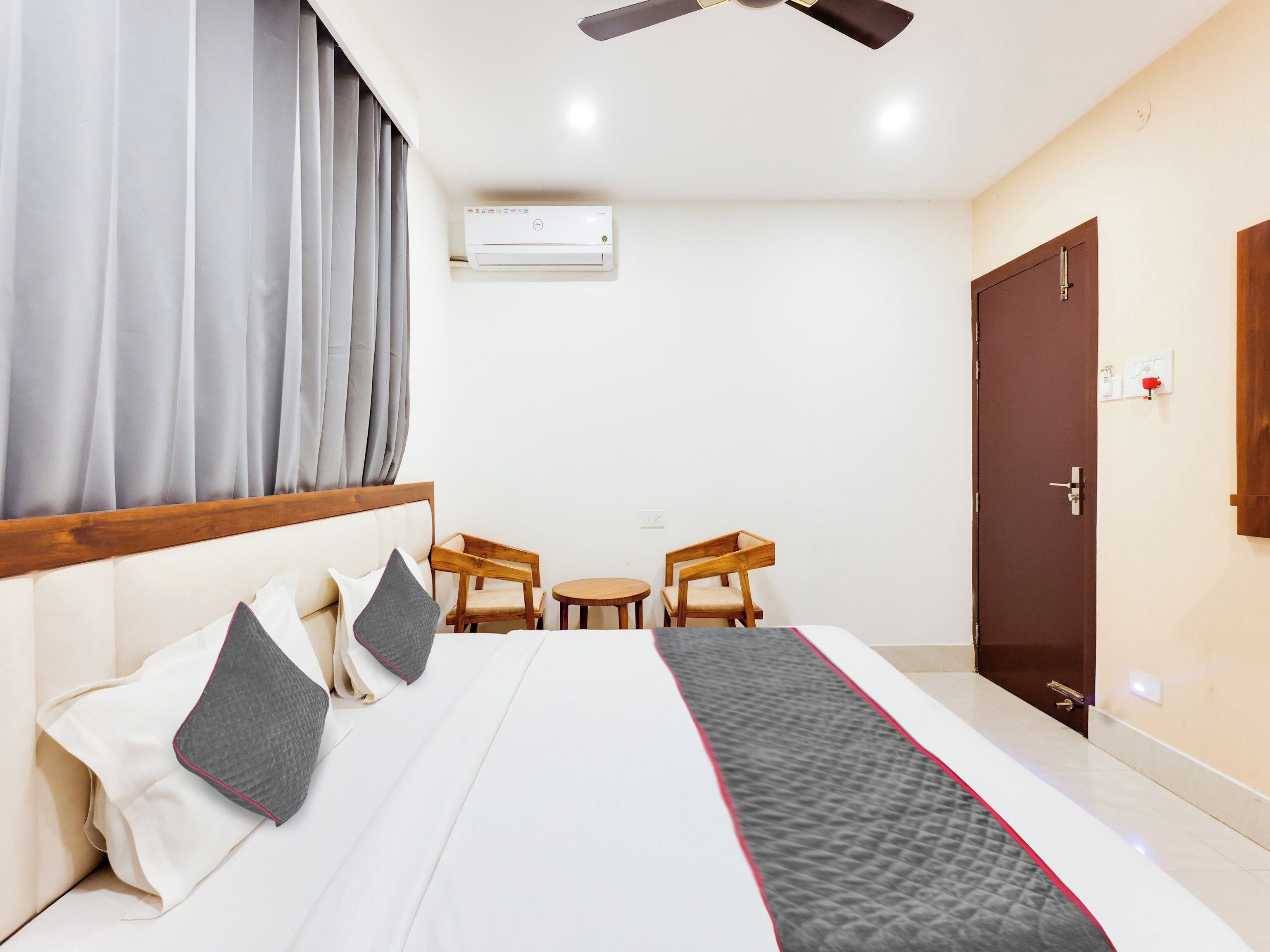 Habitación de hotel con una cama, dos sillas de madera, una mesita, un ventilador de techo, un equipo de aire acondicionado y una puerta.