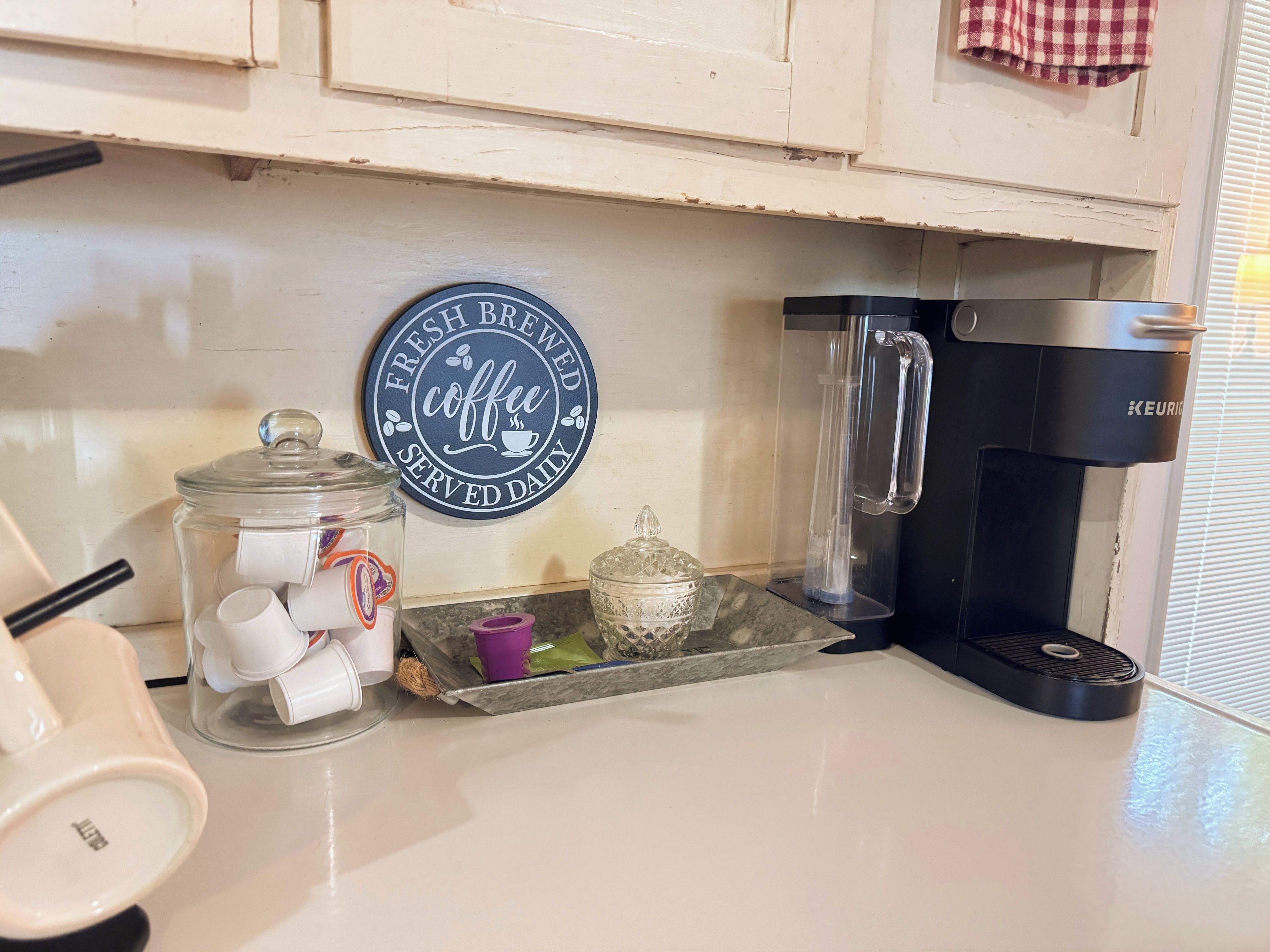 A kitchen counter with a coffee maker, a glass jar with coffee pods, and a sign that says 'Fresh Brewed Coffee Served Daily.'