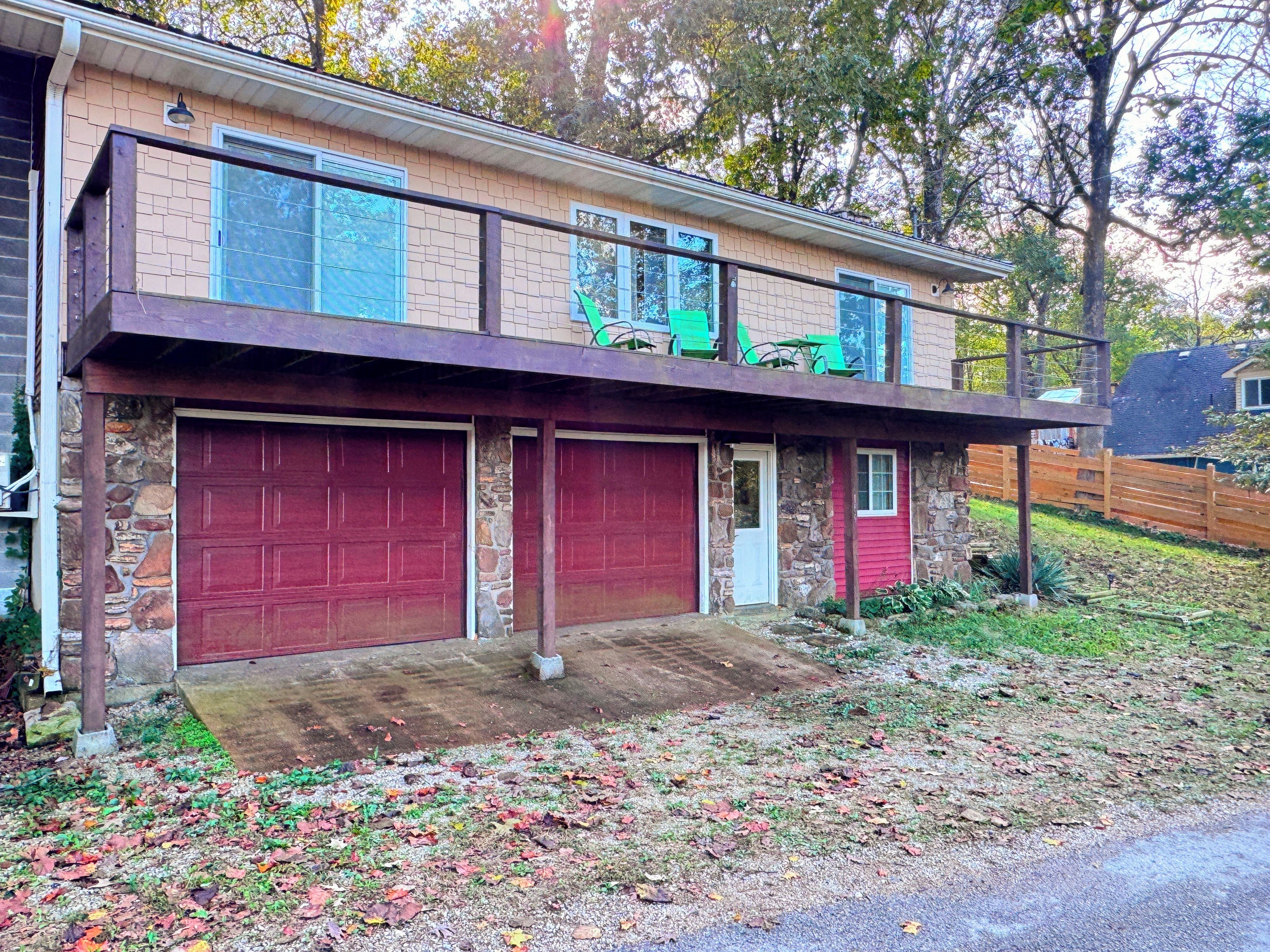 Une maison à deux étages avec une façade en pierre, une porte de garage rouge et un balcon agrémenté de chaises vertes.