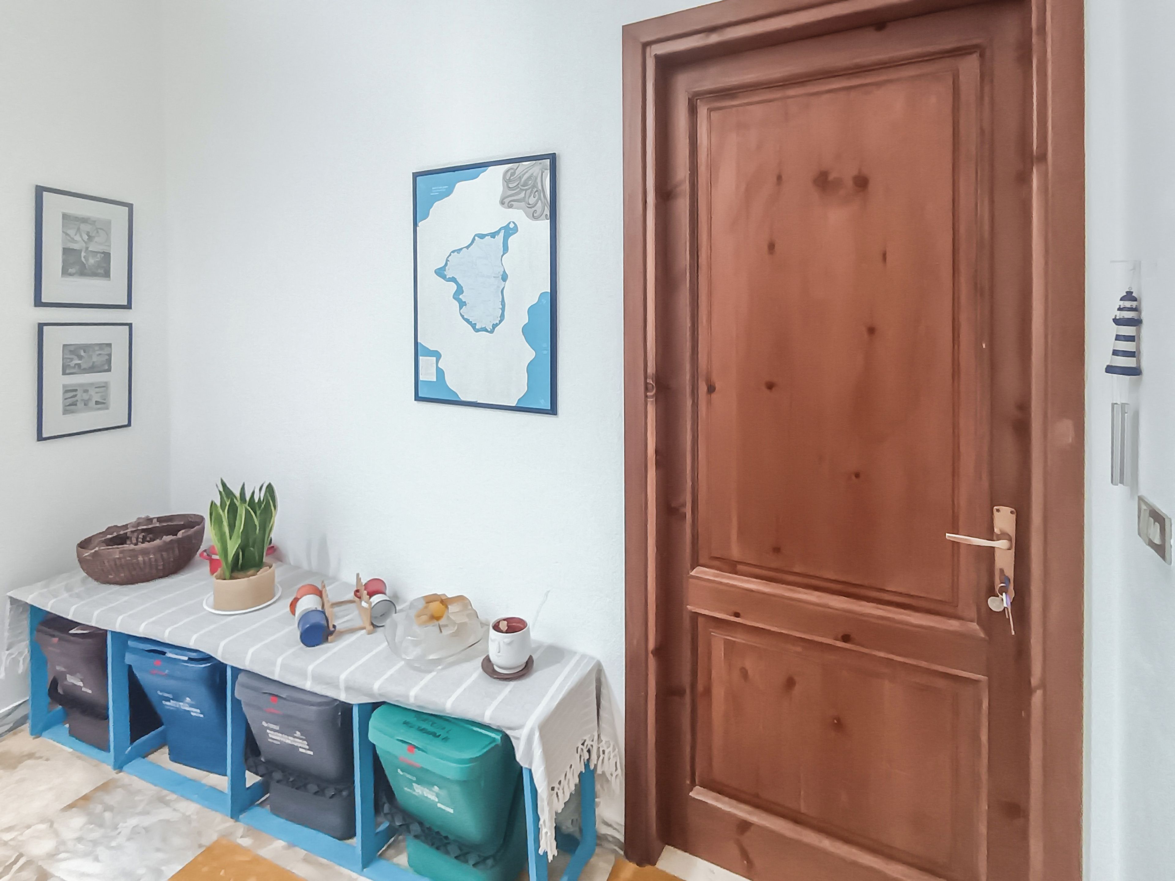 Quarto com porta de madeira, parede branca com obras de arte emolduradas, mesa listrada em azul e branco, e caixas organizadoras.