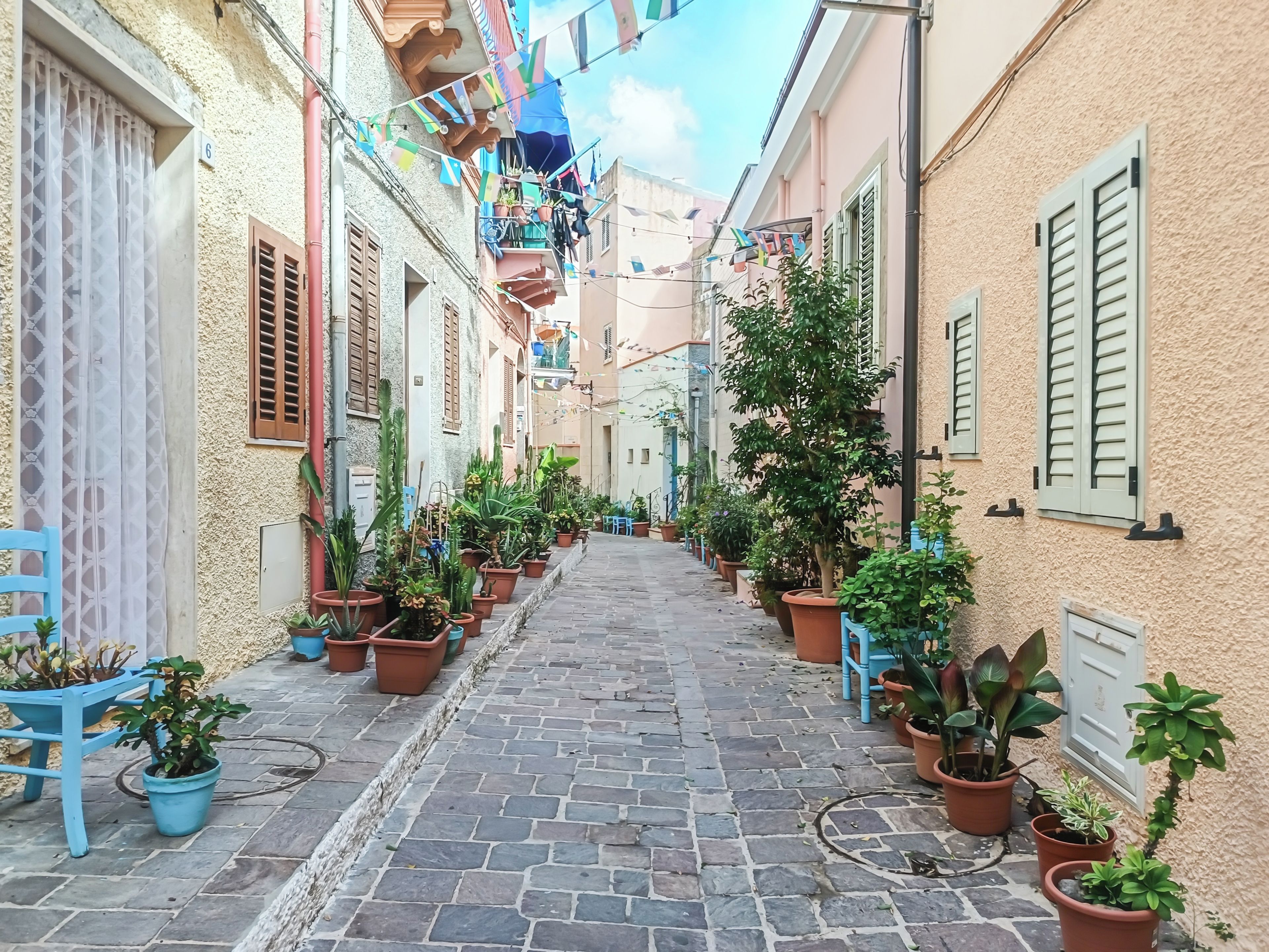 A narrow cobblestone street flanked by buildings with balconies, potted plants, and a blue chair.